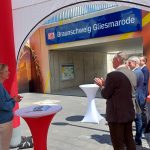 Vor der Untertunnellung der Gleise stehen ein Pavillon und einige Festtische in Tüchern, allesamt in den Bahnfarben rot und weiß. An den Tischen stehen Menschen in festlicher Kleidung und applaudieren