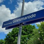 Vor grün blühenden Bäumen ragt an einem Matallpfahl ein tiefblaues Bahnhofsschild in den Himmel. Aufschrift: Gliesmarode
