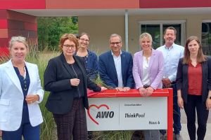 Eine Menschengruppe vor einem auffälligen Gebäude der AWO. Auf einer Tafel neben dem Eingang steht neben dem Logo der AWO Innovationslabor und ThinkPool. Das bezeichnet offenbar das moderne Gebäude aus roten Kacheln im Hintergrund. Um die Tafel am Eingang stehen einige Personen versammelt, darunter auch Julia Retzlaff und Annette Schütze.