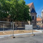 Man sieht eine augenscheinliche Baustelle mitten auf dem Domplatz (Platz der deutschen Einheit), zwischen Straße (Münzstraße) und Wasserfontänen auf dem Platz. Ein großes Viereck aus Bauzaun ist aufgebaut. Durch das Gitter sieht man einen Haufen Erde auf einer Plastikplane auf dem Boden.
