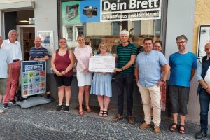 Vor einem großen Schaufenster stehen auf einem Bürgersteig eine Gruppe von Menschen. In der Mitte der Fruppe stehen Julia Retzlaff, Susanne Hahn und Heiner Hilbrich. Die drei halten einen großen Bank Check in der Hand mit einer Spende über 500 Euro.