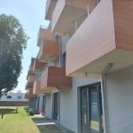 Man sieht die moderne Fassade mit bodentiefen Fenstern und Balkonen, die über die Wiese ragen