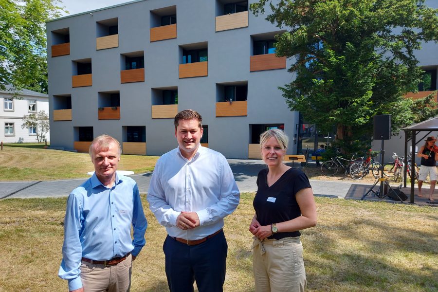 Vor dem Neubau stehen auf einer Wiese Thorsten Kornblum, Julia Retzlaff und ein Mann vom Diakonieheim in der Sonne und lächeln