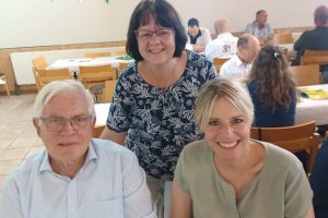 Julia Retzlaff und Gerhard Glogowski sitzen an einem Tisch und lächeln. Sonja Lerche steht hinter den beiden Stühlen und lächelt ebenfalls. Im Hintergrund sind einige Reihen von langen Tischen.