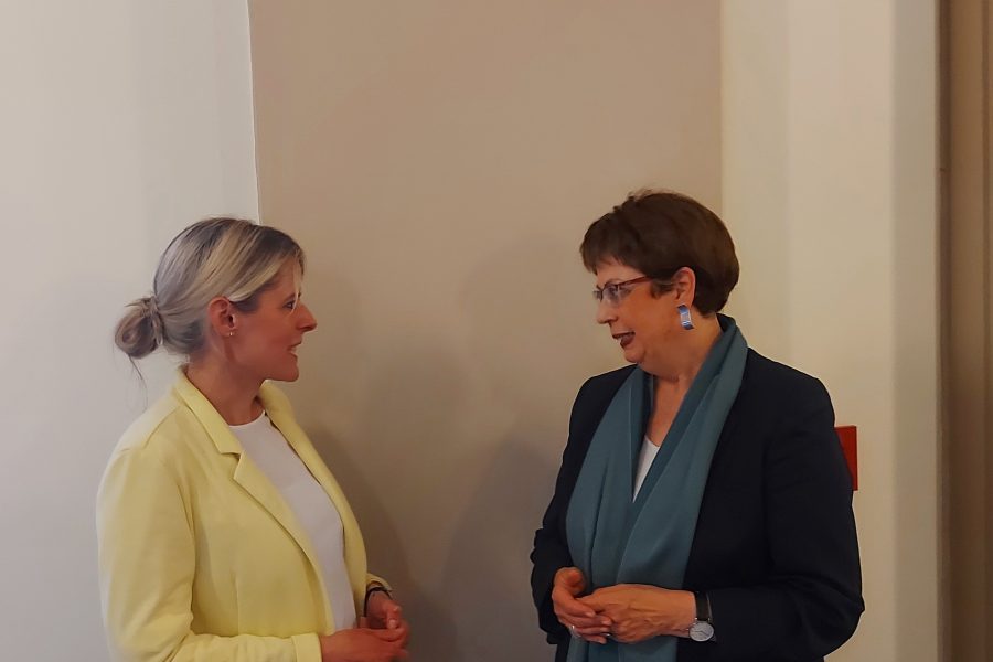 Julia Retzlaff und Birgit Hone stehen vor einer schlichten Wand im Rathaus und unterhalten sich miteinander