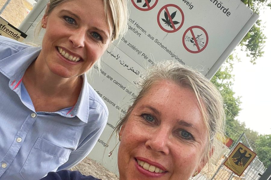 Ein Selfie (Selbstporträt, bei dem man die Kamera/das Handy auf sich selbst hält) von Annette Schütze und Julia Retzlaff vor dem Eingangsschild vor der Landesaufnahmebehörde Braunschweig
