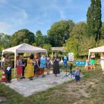 Ein Bild von der Einweihungsfeier: Vor einem der Pavillons ist eine gepflasterte Steinfläche in der Wiese, die als Bühne dient. Hier stehen dem Publikum zugewandt neun Frauen. Eine von ihnen hält ein Mikrofon