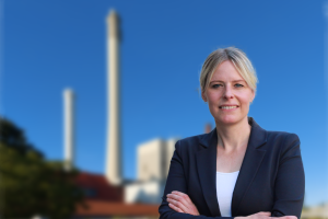 Auf dem Bild sieht man Julia Retzlaff. Sie steht mit verschränkten Armen lächelnd der betrachtenden Person zugewandt. Im Hintergrund ist verschwommen das Kraftwerk von BS Energy zu erkennen.