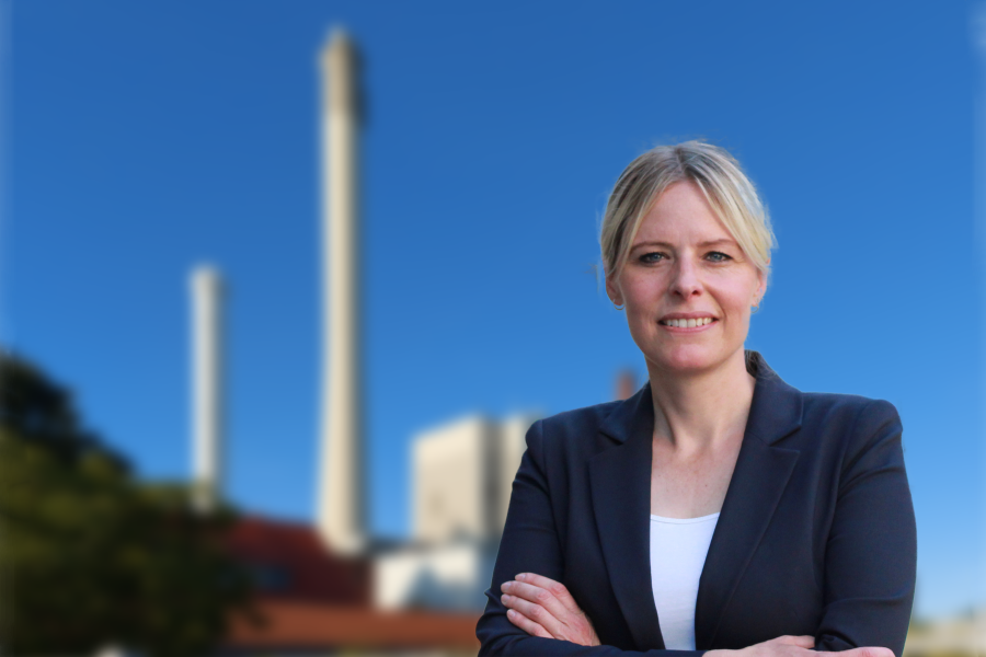 Auf dem Bild sieht man Julia Retzlaff. Sie steht mit verschränkten Armen lächelnd der betrachtenden Person zugewandt. Im Hintergrund ist verschwommen das Kraftwerk von BS Energy zu erkennen.