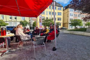 Cafe` mit Julia: Das Bild stammt vpm Andreeplatz. Zu sehen ist ein rotes SPD-Zelt mit Bistro-Tischen, Stühlen, auf denen Besucher:innen sitzen. Julia Retzlaff ist im Gespräch mit ihnen.