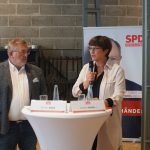 Saskia Esken spricht in ein Mikrofon. Mit ihr steht ein weiterer Podiumsgast an einem der Stehtische, der ihr aufmerksam zuhört. Im Hintergrund steht ein Stepahn Weil- Banner in der ehemaligen Fabrikhalle.