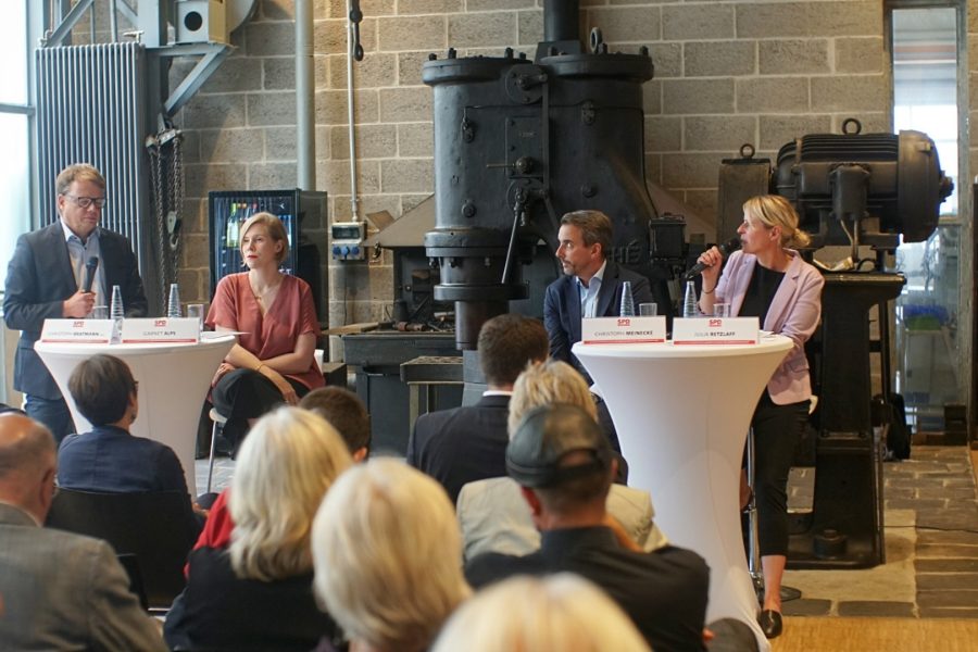 In einer ehemaligen Produktionshalle sitzen vor einem gut gefüllten Publikum Podiumsgäste auf Barstühlen an Stehtischen. Julia Retzlaff spricht ins Mikrofon. Sie und zwei weitere Gäste sind Christoph Bratmann zugewand, der bereits ein Mikrofon hält.