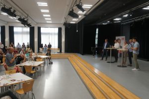 In einer Aula einer Schule ist die Veranstaltung zu sehen. Vor der Bühne stehen Tische mit je 6 Personen daran. Es ist relativ gut gefüllt. Auf der Bühne stehen hinter Stehtischen fünf Personen: Christoph Bratmann, Grant-Hendrik Tonne, Annette Schütze, Julia Retzlaff und ein Moderator.