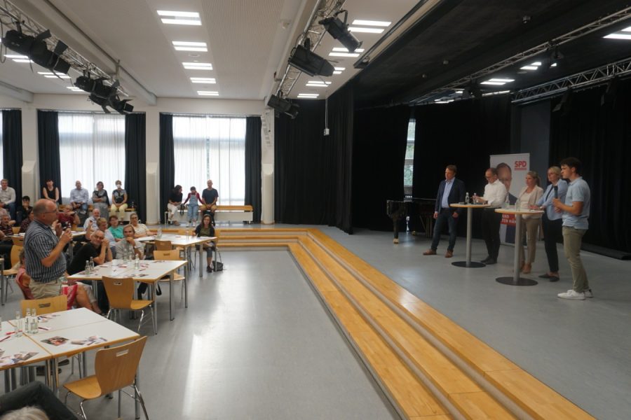 In einer Aula einer Schule ist die Veranstaltung zu sehen. Vor der Bühne stehen Tische mit je 6 Personen daran. Es ist relativ gut gefüllt. Auf der Bühne stehen hinter Stehtischen fünf Personen: Christoph Bratmann, Grant-Hendrik Tonne, Annette Schütze, Julia Retzlaff und ein Moderator.