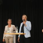 Hinter einem Stehtisch redet Julia retzlaff in ein Mikrofon. Ebenfalls im Bild auf der Bühne sind Annette Schütze, Grant Henrdik Tonne und ein Schüler vom Landesschüler:innenrat