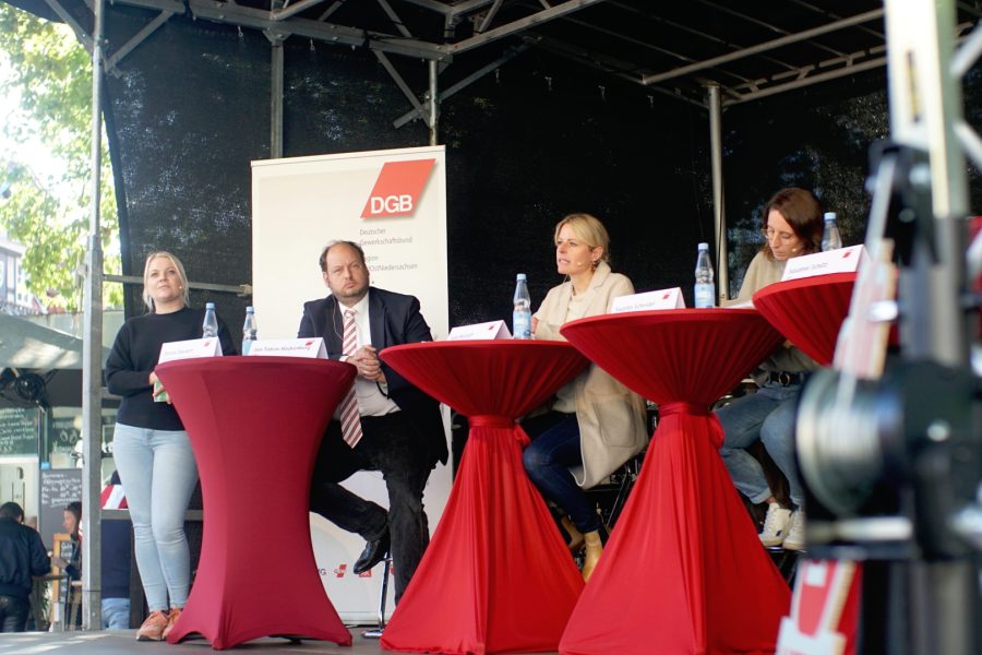 DGB Bühne auf dem Kohlmarkt. Der Bildausschnitt zeigt Modeeratorin Denise Steiner vom DGB stehend, Jan Hackenberg, CDU Kandidat an einem Stehtisch, Julia Retzlaff, SPD-Kandidatin ebenfalls an einem Stehtisch, sie spricht.